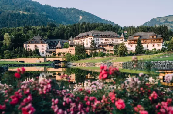 Ein luxuriöses Hotel in traditioneller alpiner Architektur befindet sich inmitten einer malerischen Landschaft. Das Gebäude spiegelt sich in einem klaren Teich, der von einer Steinbrücke überspannt wird. Im Vordergrund blühen rosa Rosen, die zusammen mit dem grünen, gepflegten Rasen und dem Hintergrund der dichten, dunkelgrünen Wälder, die Ruhe und natürliche Schönheit der Szene betonen. Die warme Beleuchtung des Hotels strahlt Gastfreundschaft und Einladung aus.