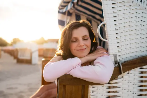Eine Frau sitzt lächelnd auf den Rand eines Strandkorbs gestützt am Strand. Die tiefstehende Sonne taucht das Bild in ein warmes Licht. 