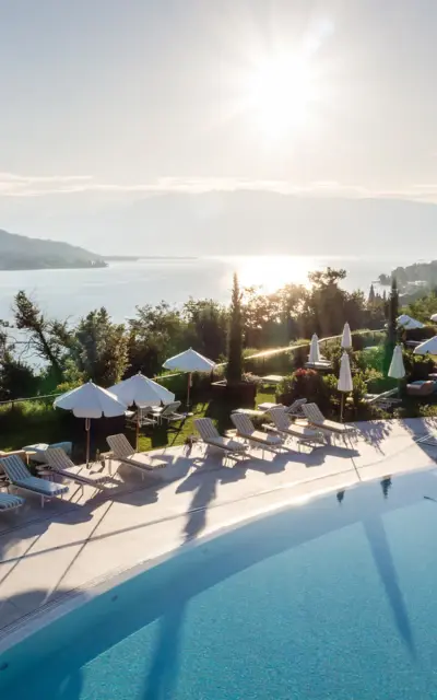 Piscina con lettini e ombrelloni sulle rive di uno specchio d'acqua, circondata da alberi e montagne.