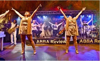 Eine Live-Performance der Band "ABBA Review" mit zwei Sängerinnen in goldenen Outfits, die mit ausgebreiteten Armen auf der Bühne stehen. Im Hintergrund spielt die Band, beleuchtet von bunten Scheinwerfern, vor einer Bühne mit dem Schriftzug "ABBA Review"