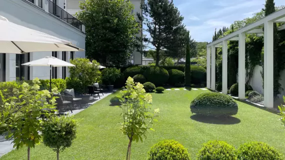 Ein gepflegter Garten mit makellosem Rasen,  der von sorgfältig beschnittenen, kugelförmigen Büschen und einigen jungen Bäumen gesäumt wird, vor einem eleganten Gebäude. Vor dem Gebäude sind weiße Sonnenschirme aufgespannt. 