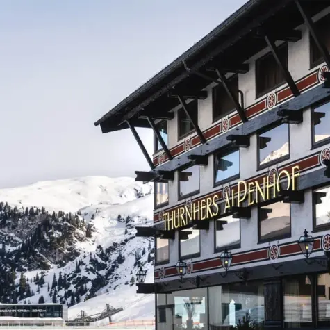 Verschneites Bergdorf bei Dämmerung, umgeben von hohen schneebedeckten Bergen. Die beleuchteten Straßen und Häuser des Dorfes erzeugen einen warmen Kontrast zur kalten, blauen Winterlandschaft.