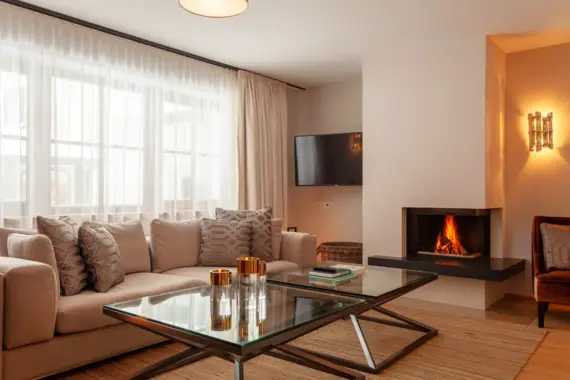 A beige sofa with lots of cushions stands in a cozy living room with a fireplace. In front of the sofa is a clas coffee table and a TV hangs on the wall in the far corner.