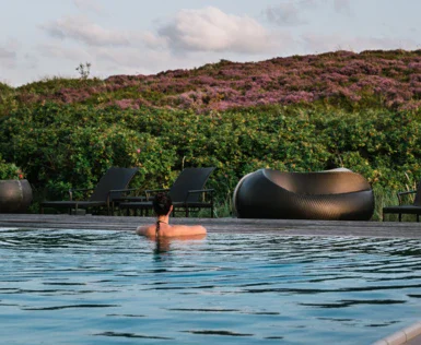 Eine Person entspannt im ruhigen Wasser eines Außenpools, umgeben von einer üppigen, blühenden Heidelandschaft. Im Hintergrund sind Liegestühle zu sehen, die entlang des Pools aufgestellt sind.