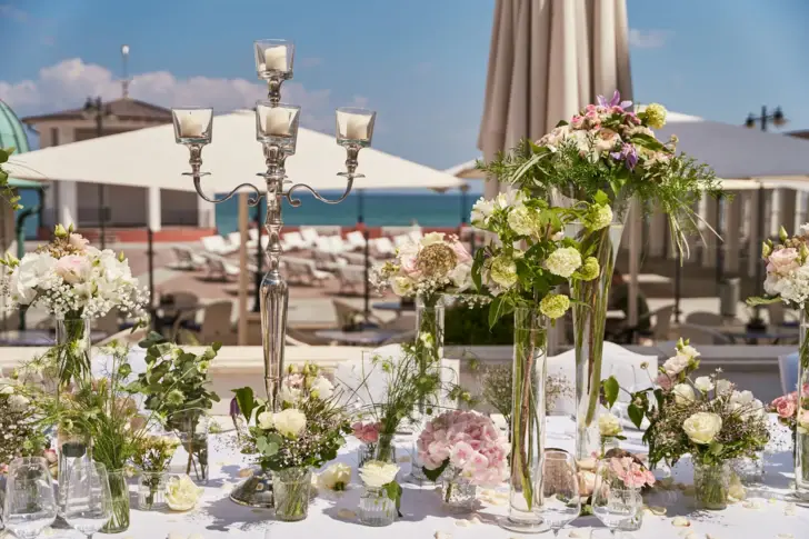 Ein Tisch im Freien, geschmückt mit einem Blumenarrangement und Kerzen als Mittelstück. Der Himmel strahlt blau und das ruhige Meer ist im Hintergrund zu sehen. 