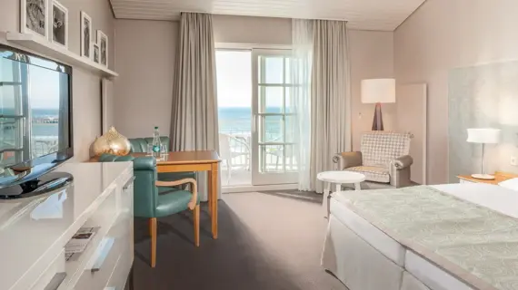 Einladendes Hotelzimmer mit Meerblick, ausgestattet mit einem Doppelbett, dessen Kopfende mit einer Tapete mit geometrischem Muster hinterlegt ist. Vor den bodentiefen Fenstern, die auf einen Balkon mit Aussicht auf die Ostsee führen, stehen schwere, graue Vorhänge und eine zarte Gardine. Eine gemütliche Sitzecke mit einem gepolsterten Sessel, ein Schreibtisch mit grünen Stühlen und ein großer Flachbildfernseher vervollständigen die moderne und komfortable Einrichtung. An der Wand über dem Schreibtisch sind Schwarz-Weiß-Fotografien arrangiert, die die elegante Atmosphäre des Raumes unterstreichen.