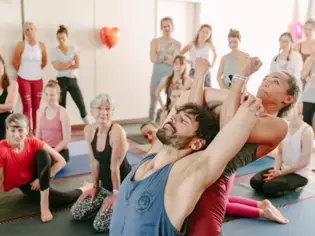 Tina Molsen hilft einem Mann in blauem Oberteil bei einer Yogaübung, während ihr im Hintergrund eine Yogagruppe aufmerksam dabei zuschaut. 