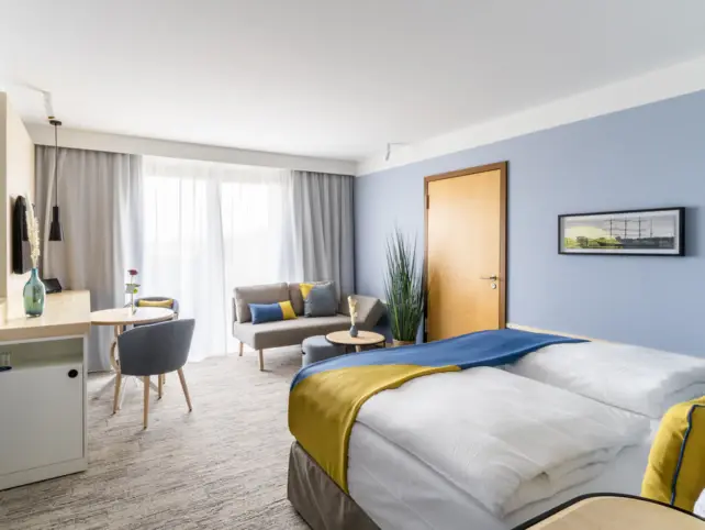 Ein modernes Hotelzimmer mit einem komfortablen Bett, dessen weiße Bettwäsche durch gelbe Akzente ergänzt wird. An der Wand gegenüber dem Bett hängt ein gerahmtes Bild. Auf der linken Seite befindet sich ein Arbeitsbereich mit einem Schreibtisch und Stuhl. Im Hintergrund ist eine Sitzecke mit einem grauen Sofa, einem runden Couchtisch und einem blauen Sessel zu sehen. 