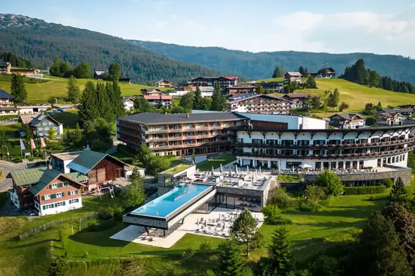 Luftaufnahme eines luxuriösen Berghotels mit moderner Architektur, umgeben von grünen Wiesen und traditionellen Alpenhäusern. Im Vordergrund ein Infinity-Pool mit Terrasse, dahinter das imposante Hauptgebäude mit Balkonen und großen Fensterfronten.