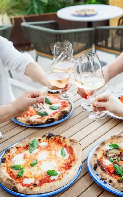 Gruppe von Personen stößt mit Weingläsern an, während sie an einem Tisch mit Pizza sitzen.