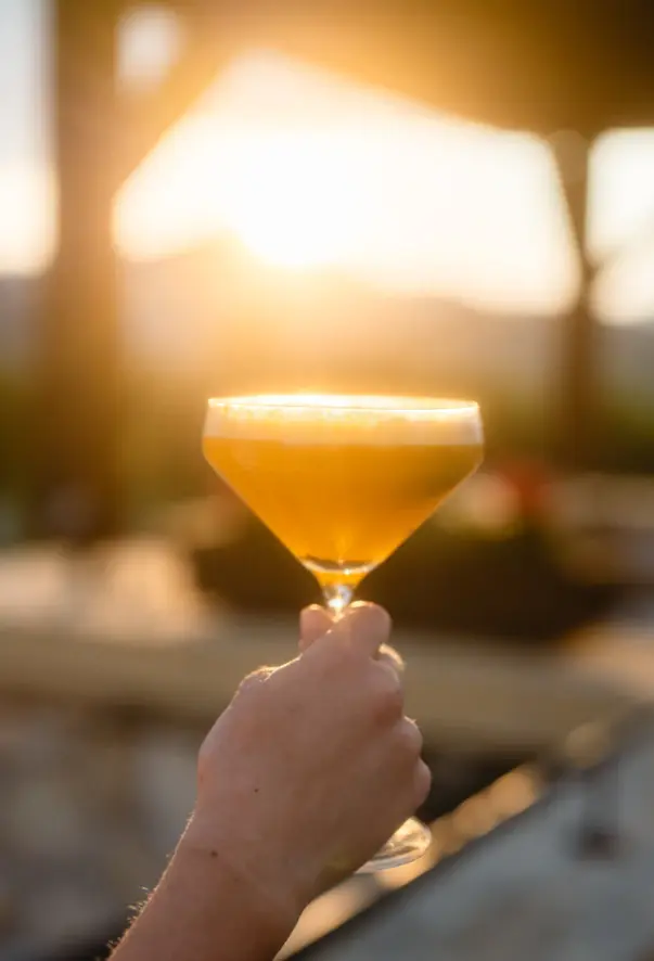 Das Bild zeigt eine Hand, die ein Glas mit einem orangefarbenen Cocktail gegen den Hintergrund einer untergehenden Sonne hält. Die Szene strahlt eine entspannte Atmosphäre aus und ist charakteristisch für einen gemütlichen Abend auf der Terrasse, vielleicht nach einem Tag voller Aktivitäten oder als ruhiger Ausklang eines warmen Tages. Die warmen Farbtöne des Sonnenuntergangs harmonieren mit dem Getränk, was ein Gefühl von Wärme und Zufriedenheit vermittelt. Das weiche Licht der Abenddämmerung und die Unschärfe im Hintergrund legen den Fokus auf das Glas und das Getränk, was den Moment als Pause und Genuss betont.