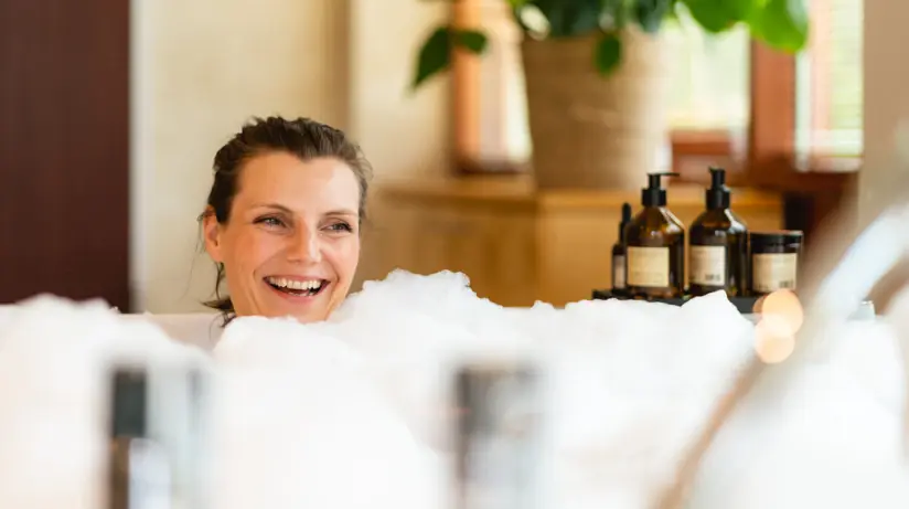 Eine Frau sitzt lächelnd, von Schaum umgeben, in einer Badewanne. Auf dem Rand sind mehrere Pumpbehälter an Pflege- und Duschsachen aufgereiht.