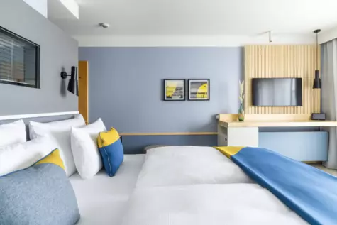 Modern eingerichtetes Deluxe Zimmer mit Meerblick im A-ROSA Travemünde. Ein Doppelbett mit weißer Bettwäsche und blauen sowie gelben Akzentkissen, an der Wand oberhalb der Betten hängen zwei gerahmte Bilder mit Strandmotiven. Gegenüber den Betten befindet sich ein großer Flachbildfernseher, umgeben von einer stilvollen Holzverkleidung. Die ruhige Farbgestaltung und das elegante Design sorgen für eine entspannende Atmosphäre mit einem Hauch von maritimem Flair.