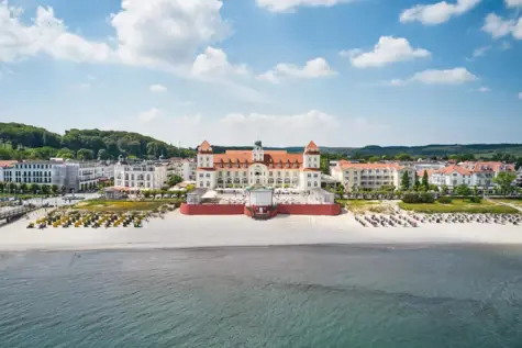 Großes Gebäude im Stil der Bäderarchitektur an einem Strand mit einem Gewässer im Vordergrund.