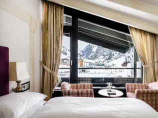 Ein Hotelzimmer mit einem Bett und zwei Sesseln und einem großen Fenster mit Blick auf eine verschneite Berglandschaft.