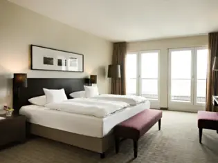 Ein Hotelzimmer mit einem großen Bett, welches mit einem dunklen Kopfteil aus Holz an einer hellen Wand steht. Darüber hängt ein länglicher Bilderrahmen, in dem drei Motive zu sehen sind.