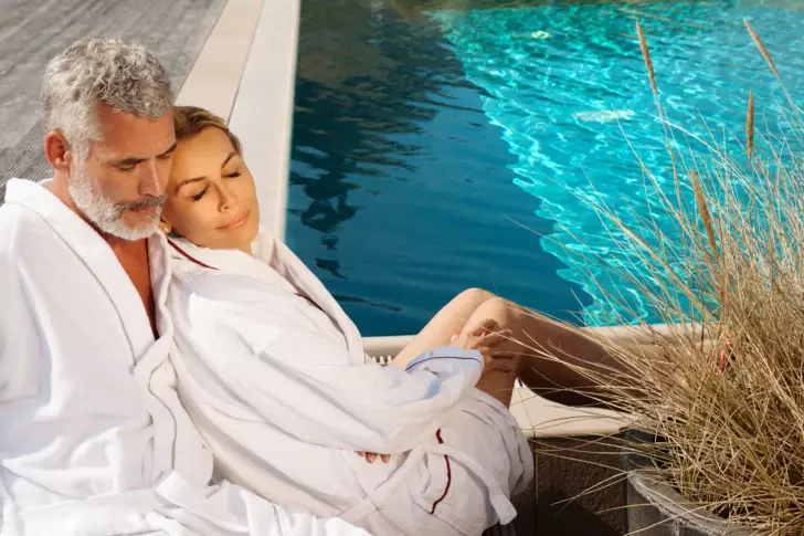 Ein entspanntes Paar in weißen Bademänteln sitzt eng umschlungen an einem Poolrand. Sie lehnen sich zurück und genießen den Moment, die Augen geschlossen.  Im Vordergrund sind zarte Gräser, die sich sanft im Wind wiegen.