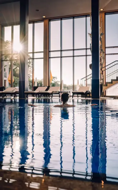Innenansicht eines Schwimmbades bei Sonnenuntergang, mit großen Panoramafenstern, durch die das warme Licht hereinströmt und sich im Wasser spiegelt. Eine Person schwimmt entspannt im Pool, umgeben von Liegestühlen im Hintergrund, was eine Atmosphäre der Ruhe und Entspannung erzeugt.