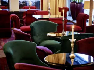 There are several smaller tables in a bar with green and red velvet armchairs.