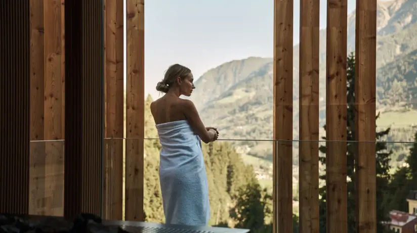 Eine Frau steht mit einem Handtuch um den Körper gewickelt auf einem Balkon und stützt die Hände auf die Glas Balustrade.