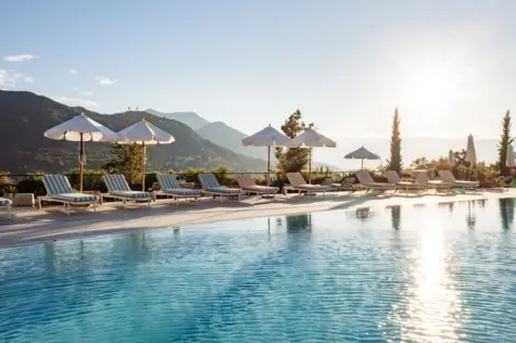 Ein ruhiger Außenpool in der Morgensonne. Um den Pool stehen gestreifte Liegen mit Sonnenschirmen und im Hintergrund sind grüne Berge zu sehen. Die Morgensonne glitzert auf der Wasseroberfläche. 