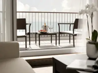 The view from a sofa out onto a terrace with two chairs and a side table. On this table is a cooler with a bottle of champagne, two champagne glasses and a bowl of strawberries. The sea can be seen in the background.
