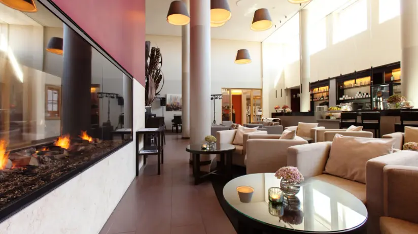 Moderne Lounge im A-ROSA Hotel auf Sylt mit gemütlichen Sitzgelegenheiten in neutralen Farbtönen, einem zentralen Kamin, der für eine warme Atmosphäre sorgt, und eleganten Pendelleuchten, die für sanftes Licht sorgen. Die stilvolle Einrichtung und das helle Ambiente laden zum Entspannen und Verweilen ein.