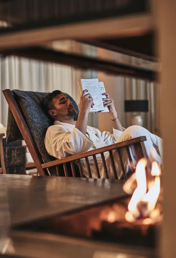 Man in a white bathrobe reads a book in an interior room. A fireplace can be seen in the foreground.