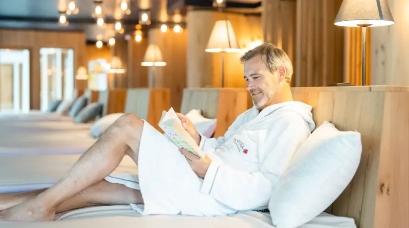 Mann in weißem Bademantel liest ein Buch auf einem Bett.