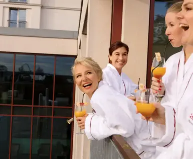 Eine Gruppe von Frauen in Bademänteln steht lachend und glücklich auf einem Balkon. Sie halten alle ein Sektglas mit Getränk in ihren Händen