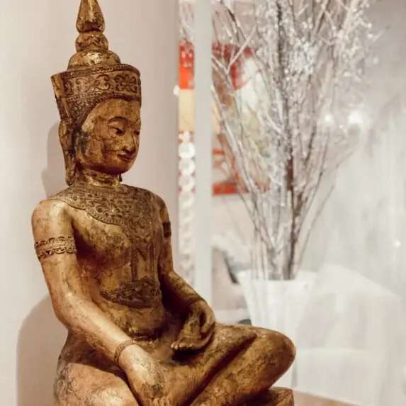 Eine goldene Buddha-Figur steht vor einer weißen Wand und im Hintergrund ist eine weiße Kunstpflanze zu sehen. 