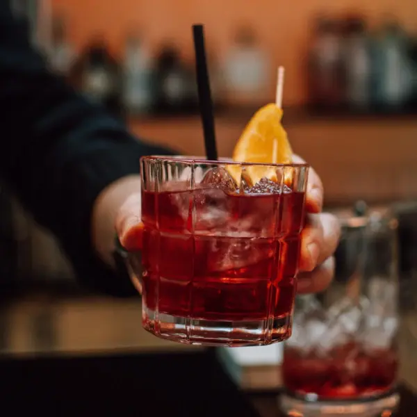Ein roter Cocktail, in einem kurzen Glas, wird von einer Hand in die Kamera gehalten. In dem Getränk befinden sich Eiswürfel, ein schwarzer Strohhalm und eine aufgespießte Orange. 