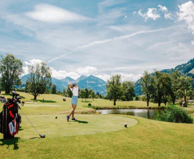 Auf dem Golfplatz von dem Hotel Kitzbühel schwingt eine Golfspielerin an einem Sommertag auf grüner Wiese mit kleinem Teich ihren Schläger. 