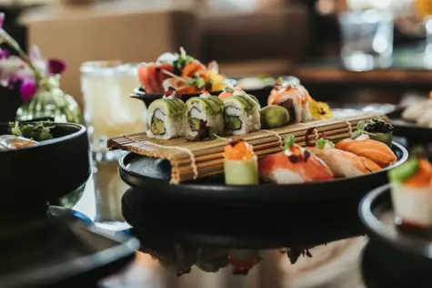  Eine Auswahl an kunstvoll arrangiertem Sushi auf einer schwarzen Platte, serviert mit Wasabi und Ingwer. Das Sushi ist im Vordergrund fokussiert, mit unscharfem Hintergrund, der das Ambiente eines Sushi-Restaurants andeutet.