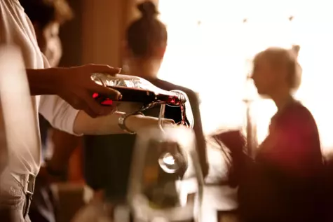 Eine Hand gießt Rotwein aus einer Glaskaraffe in ein Weinglas und im Hintergrund sind unscharf zwei Frauen zu erkenne, die einander zugewandt sind