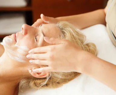 Eine Gesichtsbehandlung wird durchgeführt, bei der eine Person entspannt liegt und eine Kosmetikerin sanft eine Gesichtsmaske aufträgt.