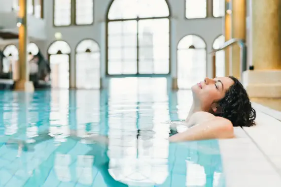 Eine entspannte Frau lehnt sich am Beckenrand eines eleganten Indoor-Pools zurück, umgeben von großen, lichtdurchfluteten Fenstern, die eine ruhige und luxuriöse Atmosphäre schaffen.