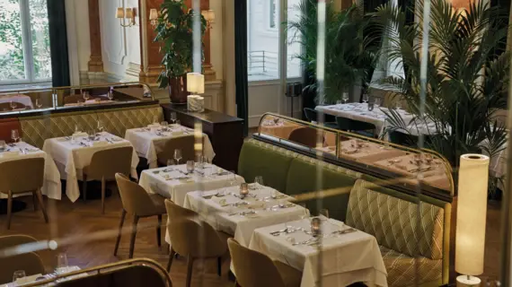 Innenansicht eines Restaurants mit Tischen und Stühlen sowie hohen Fenstern.