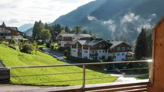 Blick von einem Balkon auf ein Bergdorf mit Bergen, Bäumen und Gebäuden im Hintergrund.