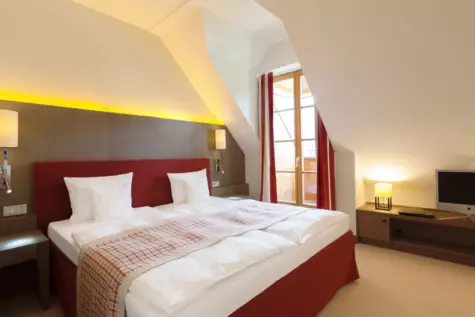 Modernes Hotelzimmer im A-ROSA Hotel Kitzbühel, ausgestattet mit einem gemütlichen Doppelbett mit rotem Kopfteil und kariertem Überwurf. Der Raum verfügt über warme Beleuchtung, die eine einladende Atmosphäre schafft, mit großen Fenstern, die auf einen Balkon führen und den Raum mit natürlichem Licht erfüllen.