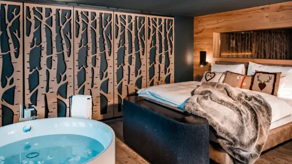 Ein Bett mit einer braunen Kucheldecke und drei Dekokissen im Alpenstil. Vor dem Bett steht eine frei stehende runde Badewanne.