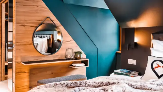 Ein Schlafbereich mit einem gemütlichen Bett im Vordergrund, auf dem eine Kuscheldecke und ein Dekokissen liegt. Hinter dem Bett ist ein Sitzhocker zu sehen, der vor einer kleinen Ablage und einem runden Spiegel steht.