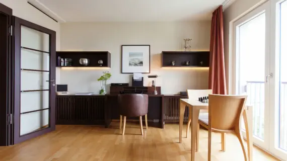 Ein Wohnbereich mit einem Schreibtisch aus dunklem Holz sowie einem kleinem hellen Tisch, der an der Fensterfront steht. 