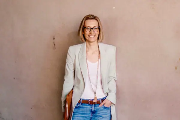 Eine Frau in Jeans steht lächelnd an einer Wand.