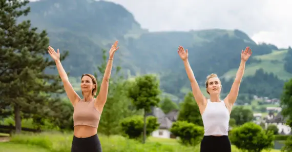 Zwei Frauen mit erhobenen Armen im Freien, lächelnd bei einer Yoga- oder Tanzpose.