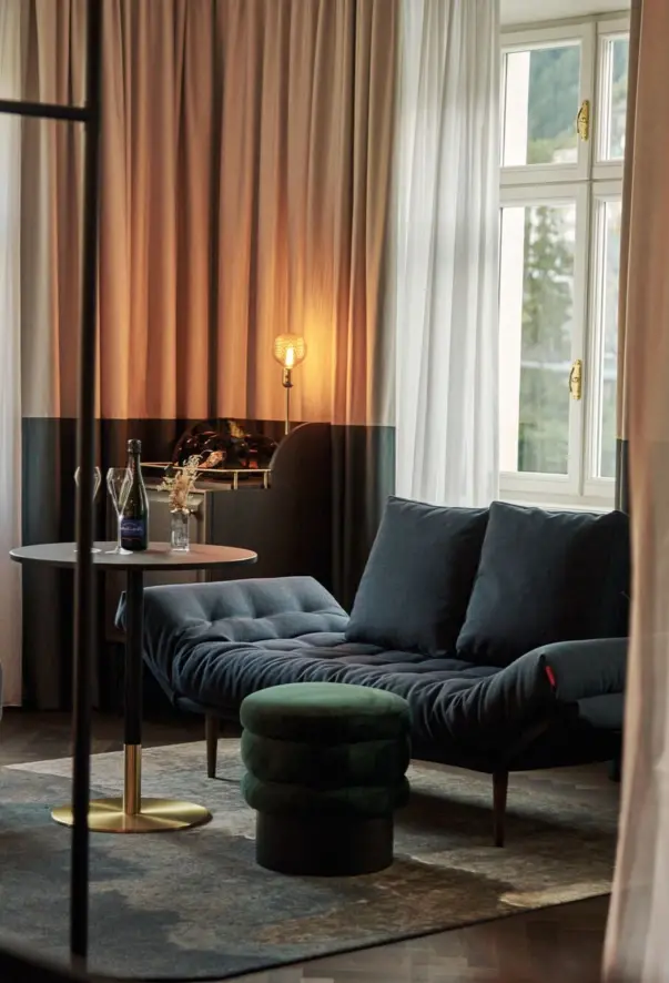 Ein Hotelzimmer mit einem blauen Sofa, einem grünen Samthocker und einem kleinen runden Tisch auf dem eine Flasche Champagner und zwei Gläser stehen.