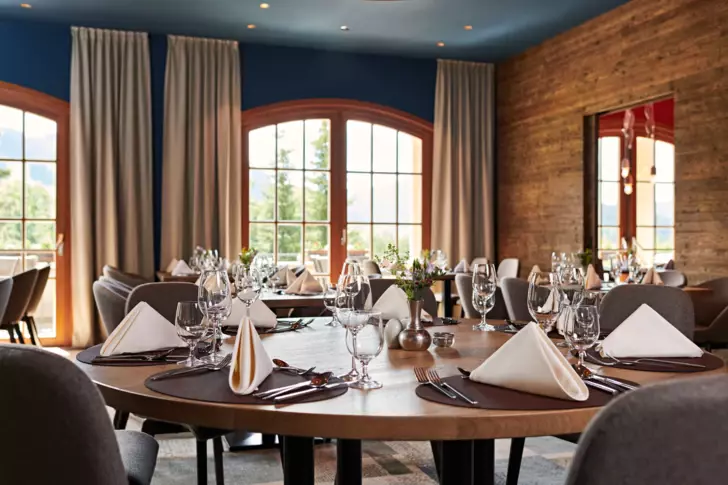 Restaurant Streif mit blauen Wänden und Holzoptik, an den großen Fenstern sind helle Vorhänge angebracht. Im Raum verteilt stehen runde, elegant gedeckte Tische auf denen in der Mitte Blumen platziert sind. 