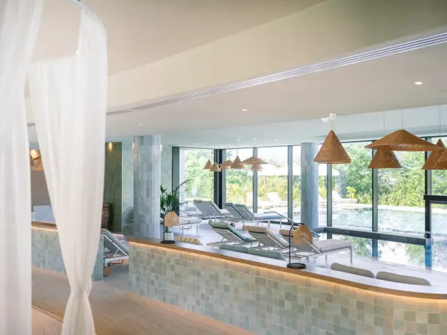 An elegantly cozy spa area with light-colored tiles and loungers that look out onto the pool through large glass windows. 