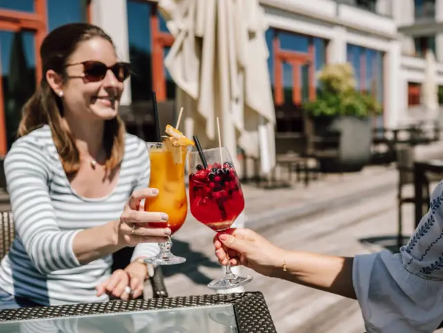 Zwei Personen genießen auf einer Terrasse die Sonne. Sie stoßen mit ihren Getränken an. Das eine Glas ist mit einem orangenen, das andere mit einem roten Getränk und Beeren gefüllt. 