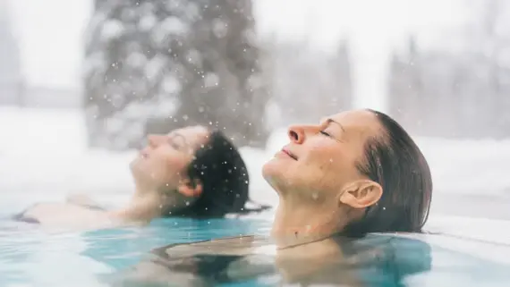 Zwei Frauen entspannen mit geschlossenen Augen im warmen Wasser eines Außenpools. Schnee fällt vom Himmel. 